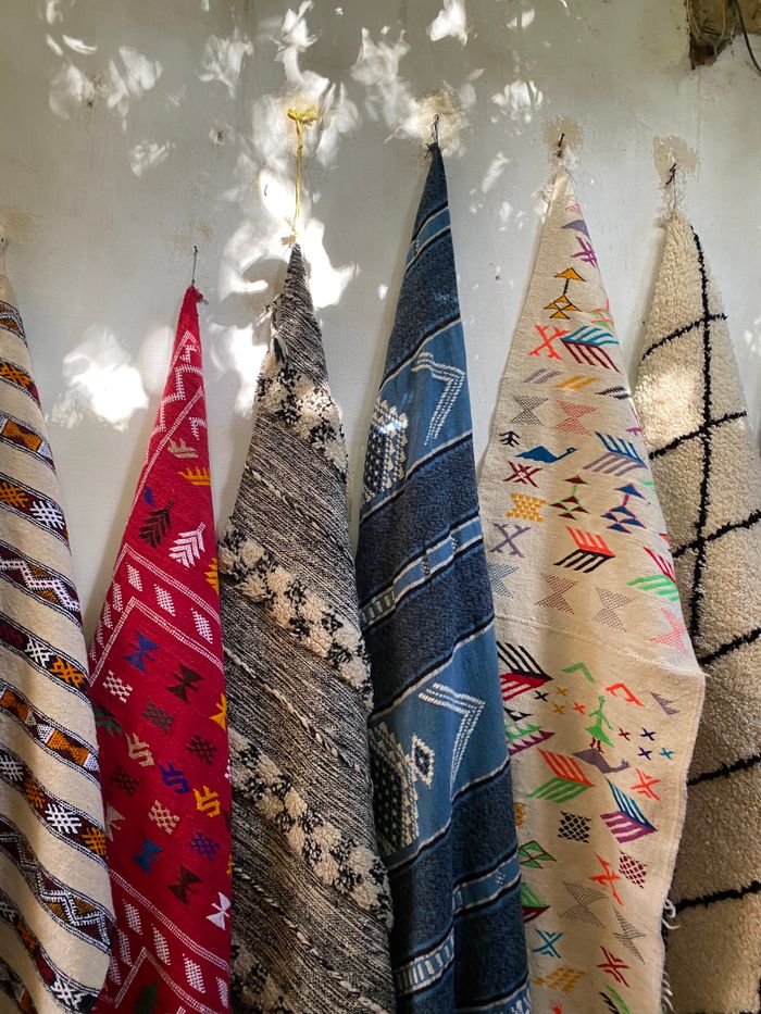 A vibrant display of traditional Moroccan rugs hanging on a wall under dappled sunlight.